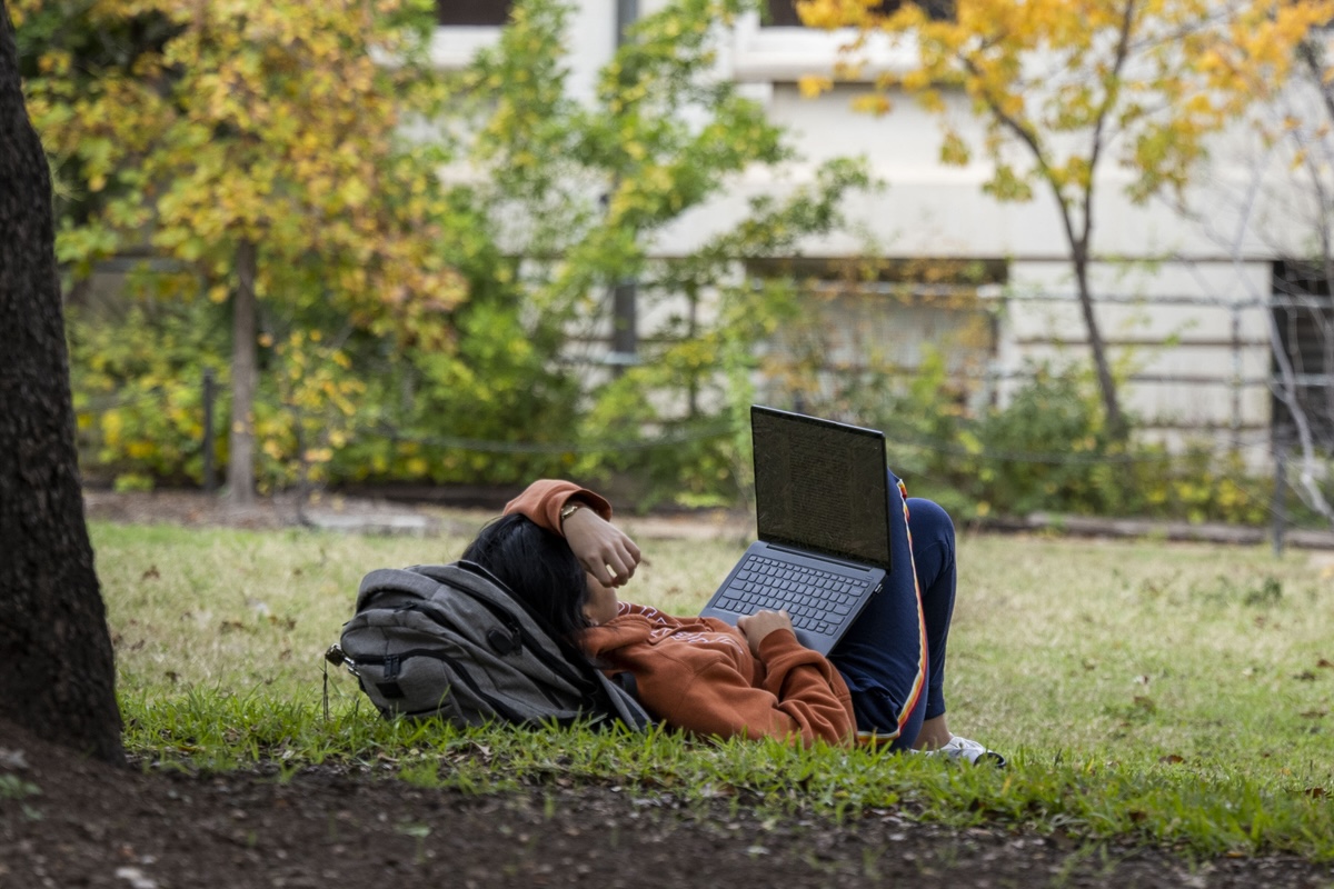 A UT student reclines against a backpack amid the trees on campus with their laptop propped open
